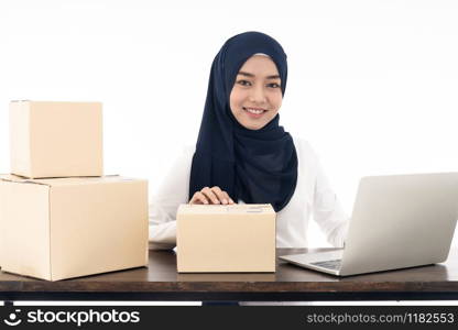 Asian muslim women merchant working with laptop computer from home on wooden table with postal parcel cardboard boxes preparing for shipping her order. Studio shot on white background using as Selling online and small business SME ideas concept