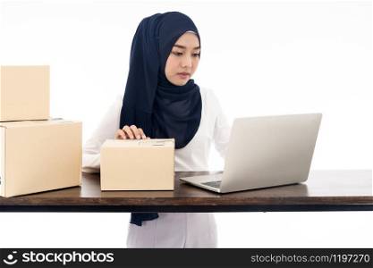 Asian muslim women merchant working with laptop computer from home on wooden table with postal parcel cardboard boxes preparing for shipping her order. Studio shot on white background using as Selling online and small business SME ideas concept