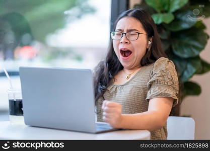 asian of freelance business female wearing wireless earphones bored yawning in front of casual working with laptop computer and coffee and smartphone in coffee shop like the background