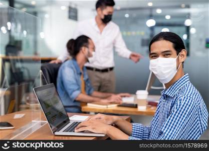 Asian Office employee with protective face mask working in new normal office with social distance practice to his colleague in background prevent coronavirus COVID-19 spreading.