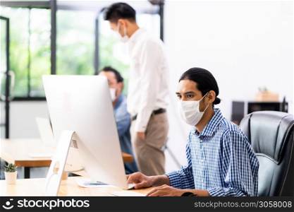 Asian Office employee with protective face mask working in new normal office with social distance practice to his colleague in background prevent coronavirus COVID-19 spreading.