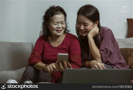 Asian old and young women online chatting by using mobile phone together in living room at home. Lifestyle, Aging, Technology and New Normal Concept.