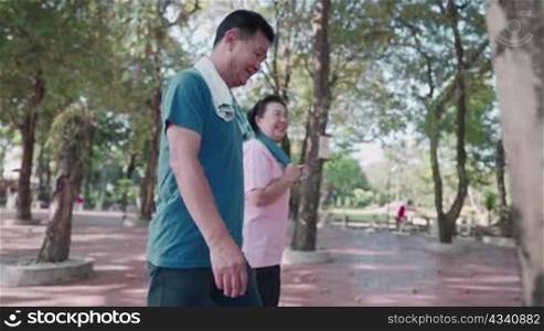 Asian old couple walking together inside public park under trees, healthy elderly retirement lifestyle, happy smiling middle age after exercising, family member outdoor activities, senior health care