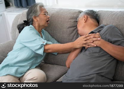 Asian old elderly man is In pain with his hands on his chest and having a heart attack in the Living room and his wife is shocked and confused, helpless. Concept of healthcare and Elderly caregivers