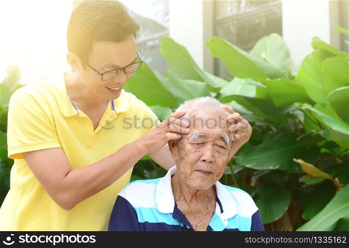 Asian old man headache, son massaging father head. Chinese family, senior retiree outdoors living lifestyle in the garden.