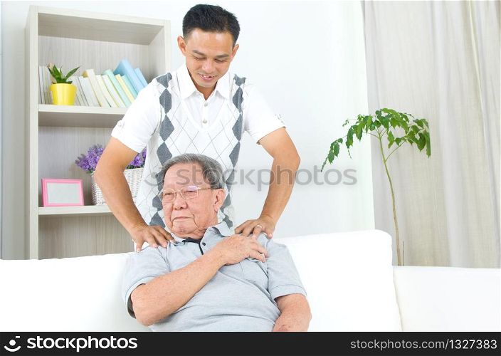 Asian old man shoulder pain, sitting on sofa , son massaging father shoulder. Chinese family, senior retiree indoors living lifestyle at home.