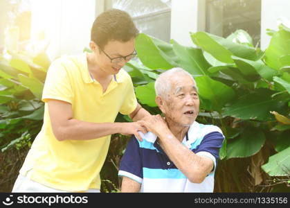 Asian old man shoulder pain, son massaging father shoulder. Chinese family, senior retiree outdoors living lifestyle in the garden.
