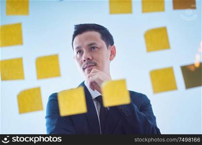 Asian pensive businessman looking at glass wall with sticker notes , brainstorming concept .