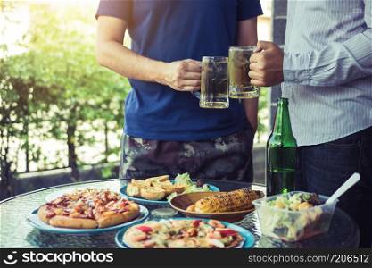 Asian people are socializing outside the house with lots of food on the table and holding a glass of beer inside clinking and enjoy together.