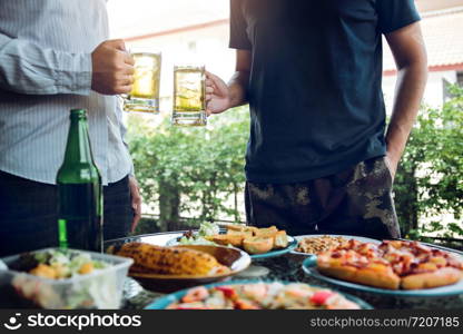 Asian people are socializing outside the house with lots of food on the table and holding a glass of beer inside clinking and enjoy together.