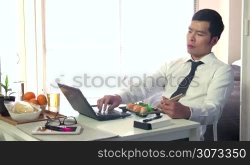 Asian people working at home with pc, young business man at work with laptop computer while having lunch and eating sushi food, businessman talking on mobile phone, busy male manager in living room