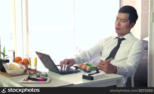 Asian people working at home with pc, young business man at work with laptop computer while having lunch and eating sushi food, businessman talking on mobile phone, busy male manager in living room