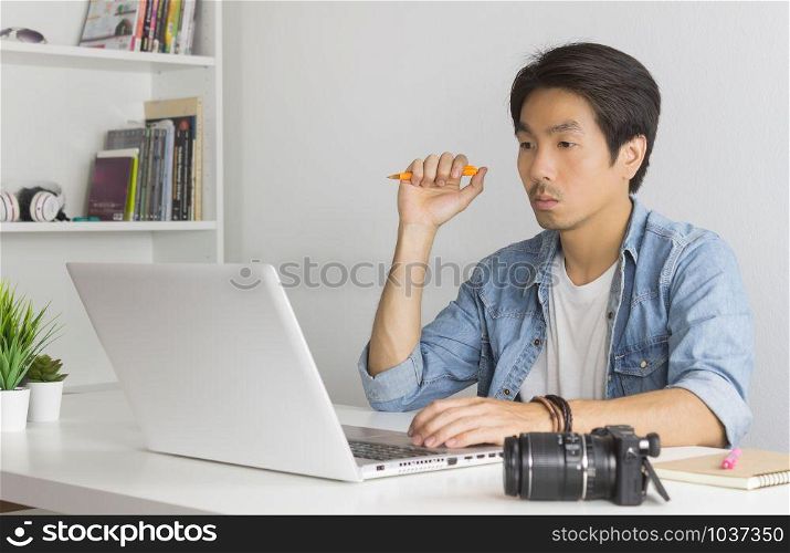 Asian Photographer or Freelancer in Denim or Jeans Shirt Hold Pen and Thinking in Front of Laptop in Home Office. Serious photographer or Freelancer working with technology