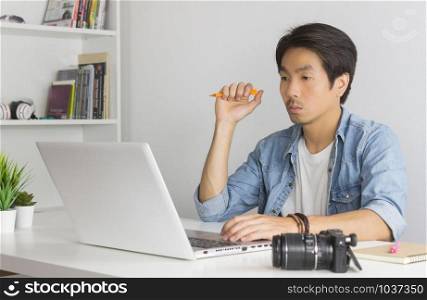 Asian Photographer or Freelancer in Denim or Jeans Shirt Hold Pen and Thinking in Front of Laptop in Home Office. Serious photographer or Freelancer working with technology