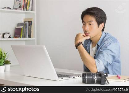 Asian Photographer or Freelancer in Denim or Jeans Shirt Serious Thinking in front of Laptop in Home Office. Photographer or freelancer working with technology