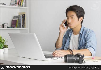 Asian Photographer or Freelancer in Denim or Jeans Shirt Talking with Customer by Smartphone in front of Laptop in Home Office. Photographer or freelancer working with technology