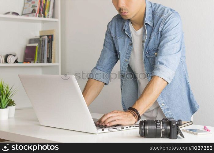 Asian Photographer or Freelancer in Denim or Jeans Shirt Working with Laptop in Standing Posture in Home Office. Photographer or freelancer working with technology