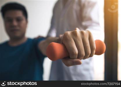 Asian physiotherapist helping a patient lifting dumbbells work through his recovery with weights in clinic room.