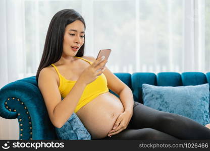 Asian Pregnant holds hands on belly and using the smart mobile phone on the sofa in modern house,preparing to be motherhood with good healthy