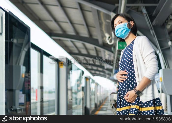 Asian Pregnant Woman in face mask waiting commuter skytrain for traveling to work. Young mother life in public transportation during pandemic virus. Health care in New normal and social distancing.
