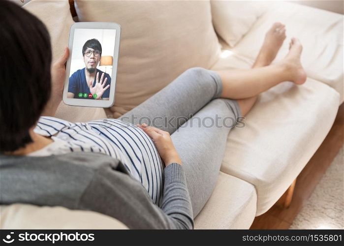 Asian pregnant woman lying on sofa making video call with digital tablet. Happy Young mother enjoying internet technology with her husband at home.