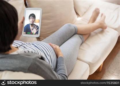 Asian pregnant woman lying on sofa making video call with digital tablet. Happy Young mother enjoying internet technology with her husband at home.