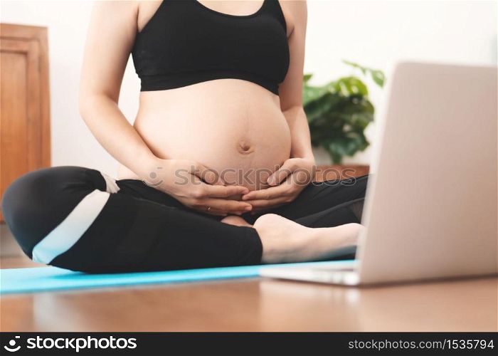 Asian Pregnant Woman practicing yoga at home with online video training class on laptop, Female in sportswear touching her belly and doing exercises during pregnancy, healthy lifestyle