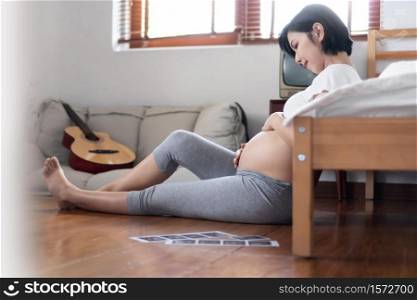 Asian Pregnant woman sitting on floor near her bed and touching her belly in bedroom. Beautiful Young Female looking and hugging her tummy at home. Tenderness, Motherhood, Pregnancy