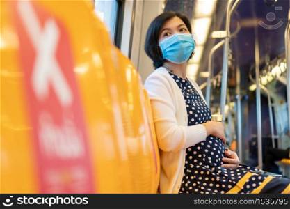 Asian Pregnant Woman wear face mask touch her belly and sit on yellow seat in sky train while go to office. Businesswoman with social distance while pandemic covid-19 or coronavirus. New normal life