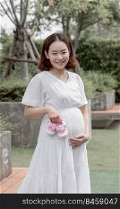 Asian prenant beautiful mother holding pink girl&rsquo;s shoes while standing in garden