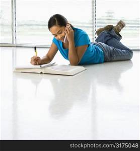 Asian preteen girl lying on floor doing homework while talking on cell phone.