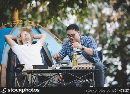 Asian relaxed man using smartphone and waiting for his friend cooking with tent background in c&ing. Cooking set front ground. Outdoor cooking, traveling, c&ing, lifestyle concept.
