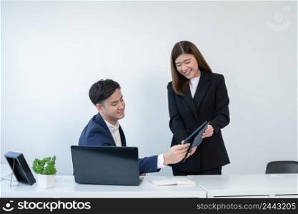Asian secretary woman is holding and showing company performance report on tablet to her young handsome boss at office with a smiling face. Business people working together with smart online devices