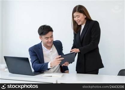 Asian secretary woman is holding and showing company performance report on tablet to her young handsome boss at office with a smiling face. Business people working together with smart online devices