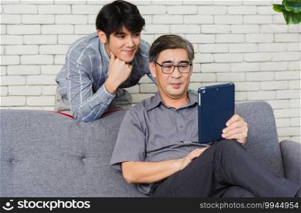 Asian senior businessman with digital tablet discuss together with young team in office. Father man and his son sit on sofa talking chatting on video call conference on tablet in living room at home