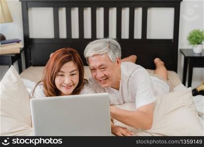 Asian senior couple using laptop at home. Asian Senior Chinese grandparents, husband and wife happy after wake up, watching movie lying on bed in bedroom at home in the morning concept.