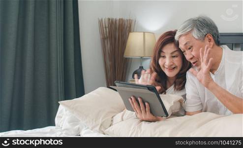Asian senior couple using tablet at home. Asian Senior Chinese grandparents, video call talking with family grandchild kids while lying on bed in bedroom at home in the morning concept.