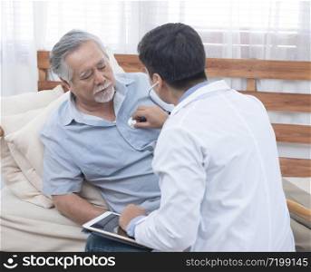 Asian senior elderly old man with mask on lying on sofa while young caucasian doctor sit on knee check his heart beat and old senior woman sit beside with care and worry .healthcare concept.
