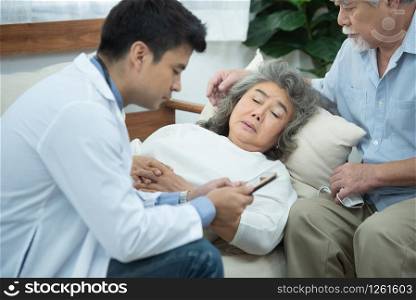 Asian senior elderly old patient woman laying down on sofa in home waiting for result from smart young caucasian doctor smartphone while check up in home with grandfather take care beside.