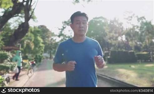 Asian senior man running inside the running track at the park on a sunny day in slow motion. Retirement lifestyle activity. Health care motivation concept, endurance cardio exercise, front view