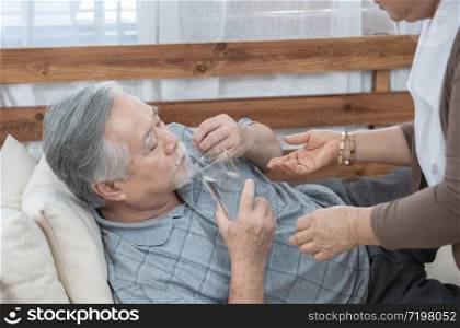 Asian Senior man taking medicines and drinking water while lie on couch. Old woman take care his husband while his illness at the house.