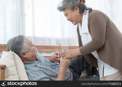 Asian Senior man taking medicines and drinking water while lie on couch. Old woman take care his husband while his illness at the house.