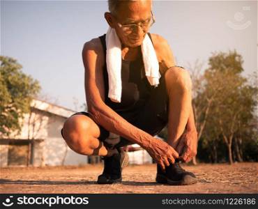 Asian senior man tying his laces of running shoes to prepare jogging in the park for good health. Healthcare concept.