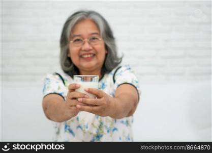 Asian Senior woman holding glass milk while relaxing on a sofa living room for retirement wellness. Elderly Woman drinking a glass of milk to maintain her wellbeing. Concept of wellness and healthy.