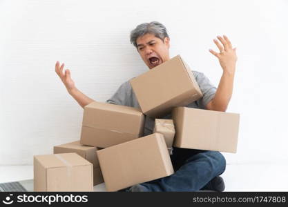 Asian Shopaholic man Shocked and sitting on the floor in the living room and Cardboard Box on top of him after The courier delivered to the home. Concept of shopping online and Shopaholic.