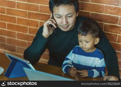 Asian single dad with son are looking the cartoon via technology laptop together with phone calling for work when living in loft house for Self learning or home school,Family and single father concept
