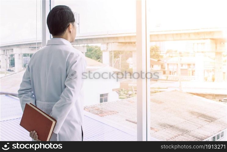 Asian smart doctor wearing eyeglasses, holding books while standing beside window. Education and Career concept.