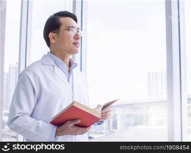 Asian smart doctor wearing eyeglasses, studying and reading books while standing beside window. Education and Career concept.