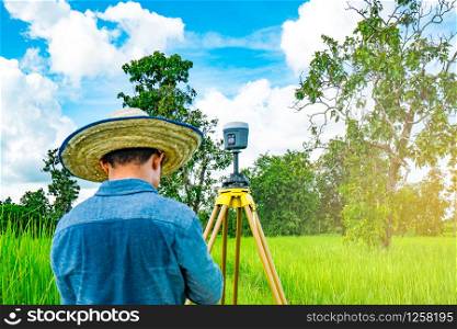 Asian smart engineer or surveyor in black jeans and long sleeve shirt and woven bamboo hat. He is working on controller screen for surveying land in rice field, Thailand. GPS surveying instrument.