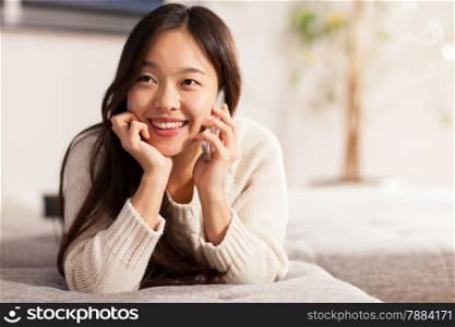 Asian smiling woman is phoning while lying on the sofa at home